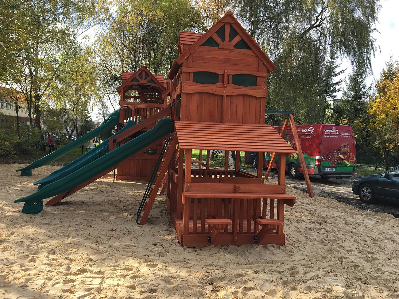 Wooden playground structure with slides and sand area, trees and vehicles in the background